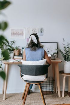 Back view of anonymous female sitting at wooden table with netbook and notebook while working in light room with potted plants