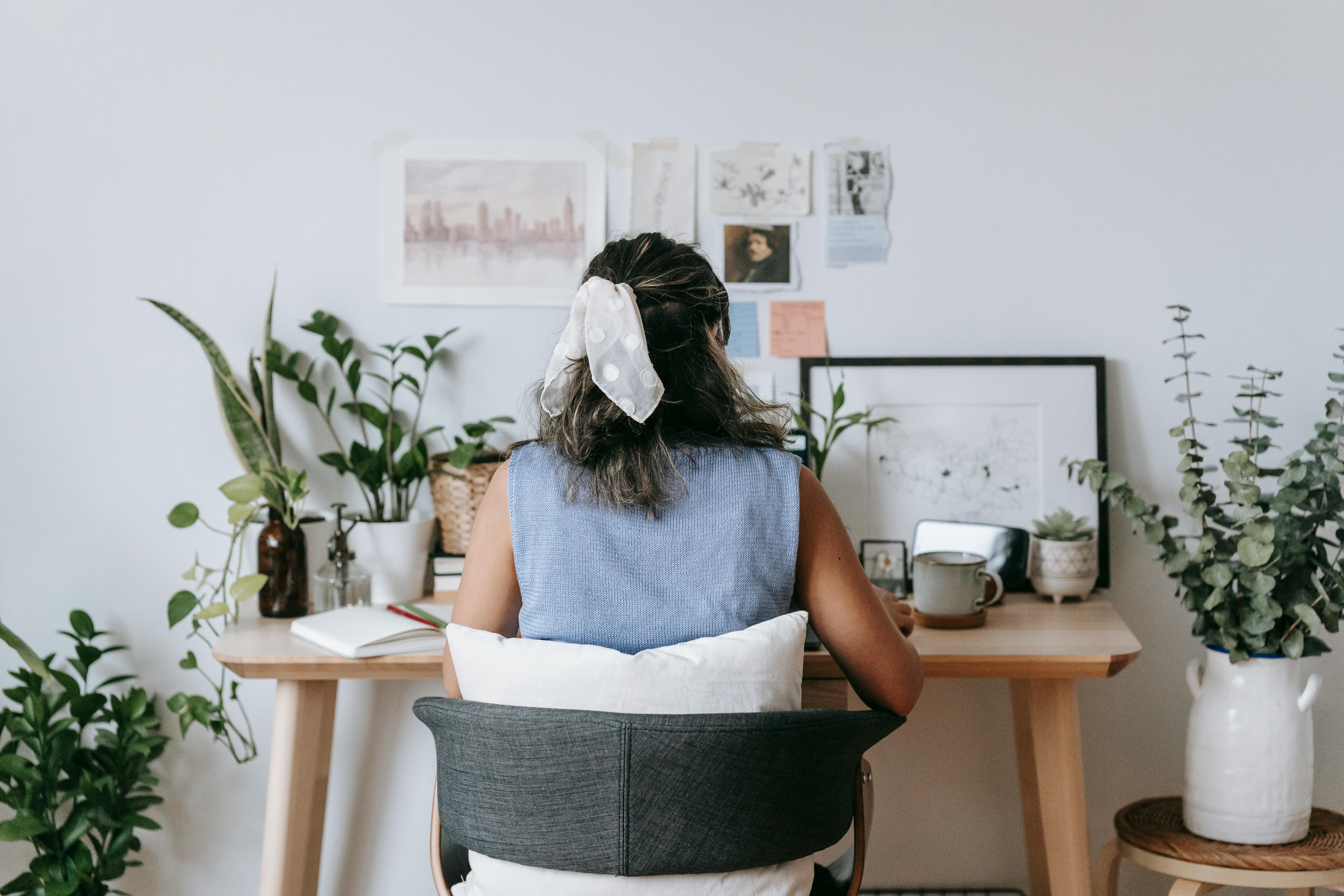 Back view of unrecognizable female florist sitting in armchair at table with assorted potted plants and opened notepad