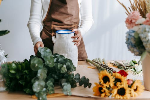 Crop anonymous female florist in apron taking green branch placed near sunflowers while working in floral shop