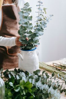 Crop unrecognizable woman in apron against desk with evergreen plant sprigs in vase on light background