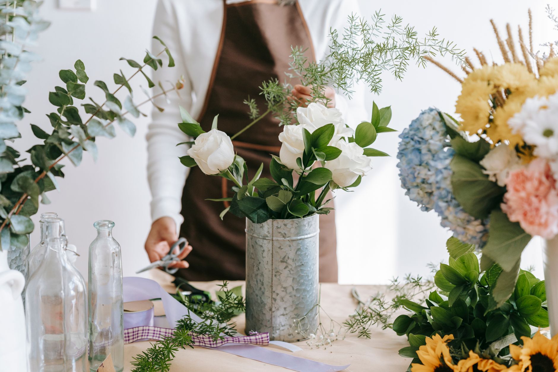 Crop unrecognizable person with Asparagus plant sprig against blooming roses and Ruscus in vase on table in shop