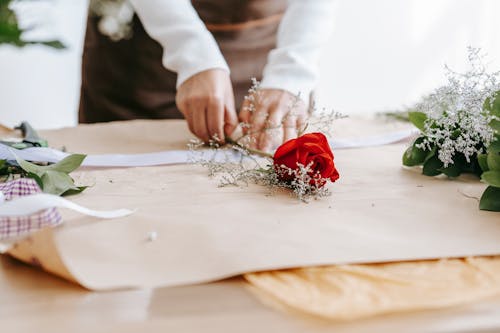 Crop anonymous person composing bright red rose and branches with delicate white flowers on table
