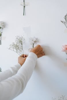An unrecognizable person arranges delicate flowers on a white wall, creating a minimalist floral display.