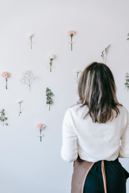 Free Back view of anonymous woman in apron looking at composition of flowers on white wall Stock Photo