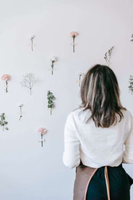 Back view of anonymous woman in apron looking at composition of flowers on white wall