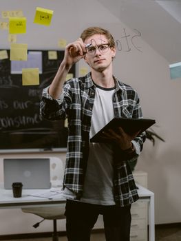 A young man with glasses writes on a glass panel in a modern office setting, holding documents.