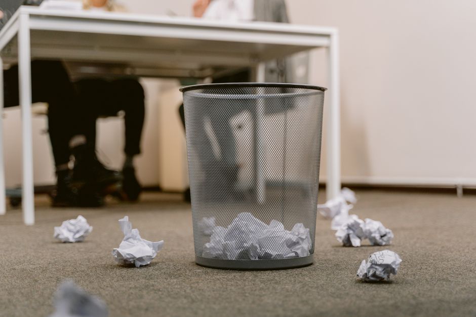 Office floor with a visible trash bin and crumpled papers scattered around, blurring human figures.
