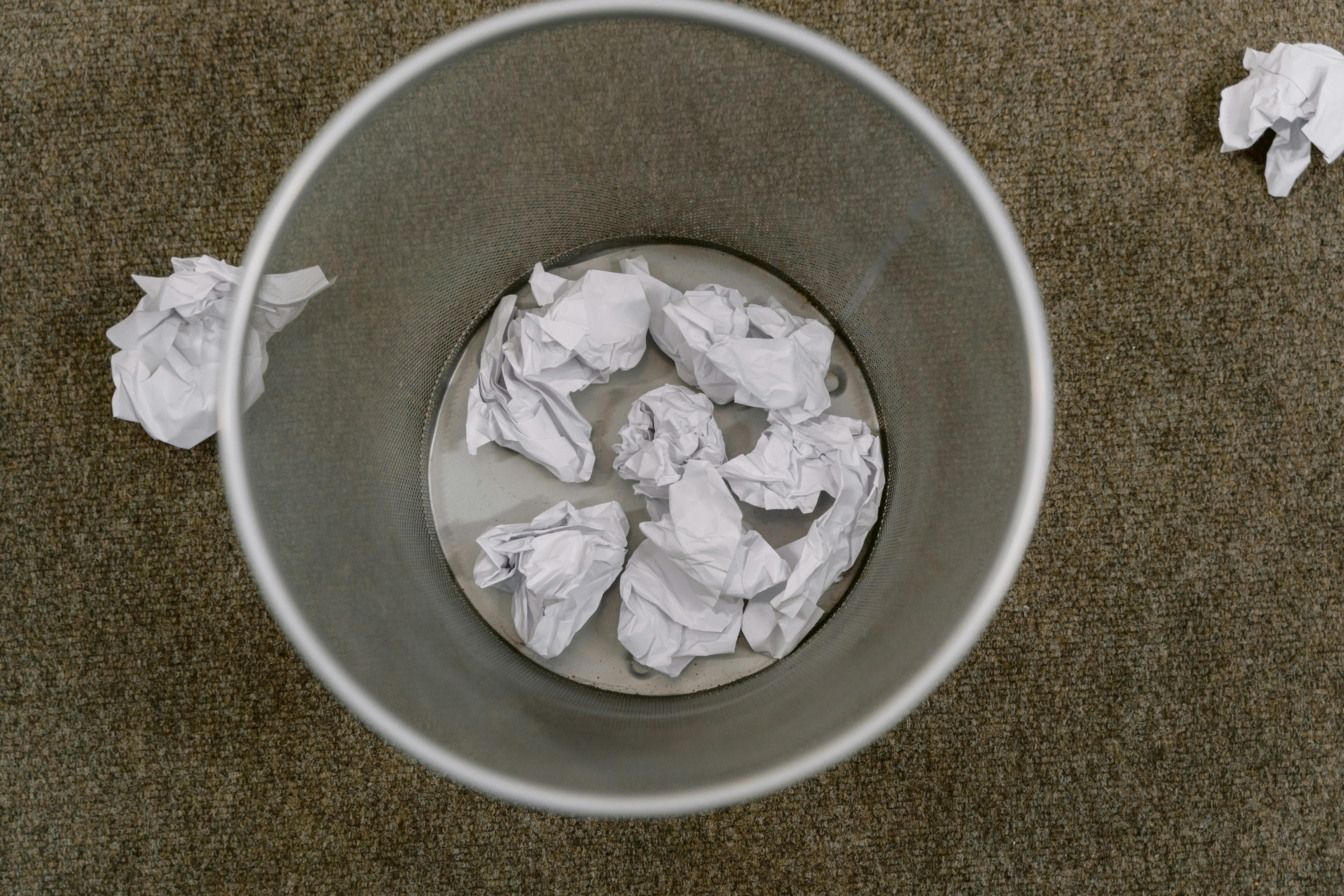 Overhead view of a trash can filled with crumpled paper on a carpeted floor.