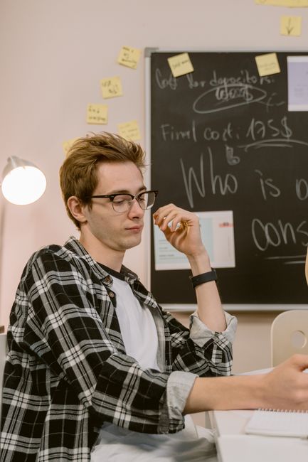 Young man wearing glasses and plaid shirt sitting at a desk in an office setting, focused on work. Young man wearing glasses and plaid shirt sitting at a desk in an office setting, focused on work.