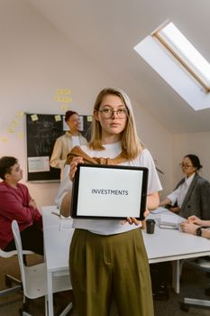 Young woman holding a tablet with 'INVESTMENTS' text in a modern office setting.