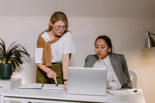 Two women working together in a modern office, collaborating on a project using a laptop.