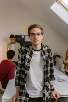 A young man wearing glasses stands confidently in a modern office setting.