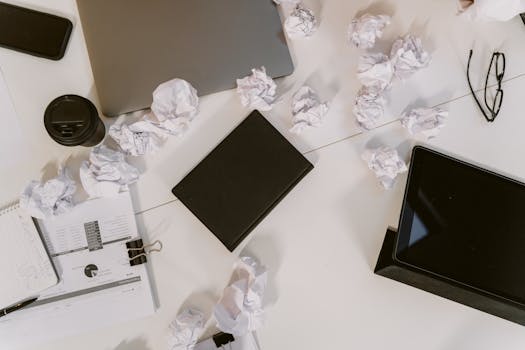 Top view of a cluttered office desk with crumpled papers, laptop, and eyeglasses.