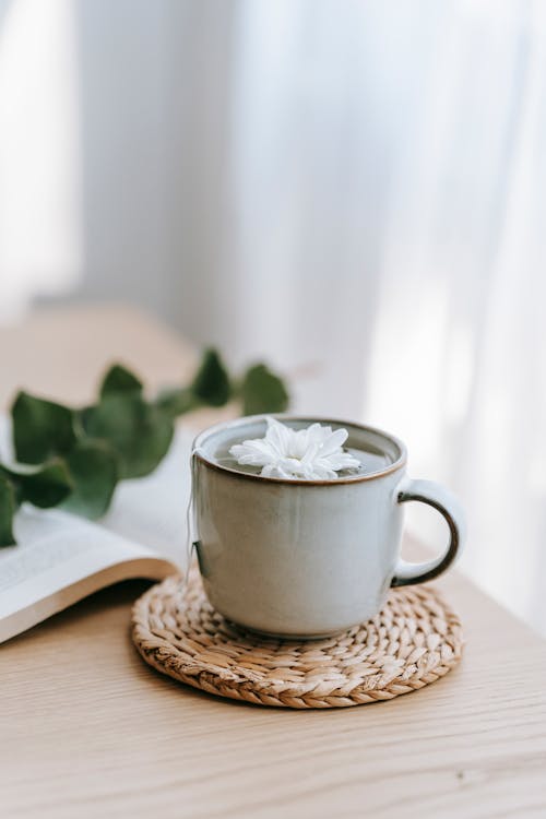 Cup of green hot beverage with blossoming white flower on straw mat against book in house