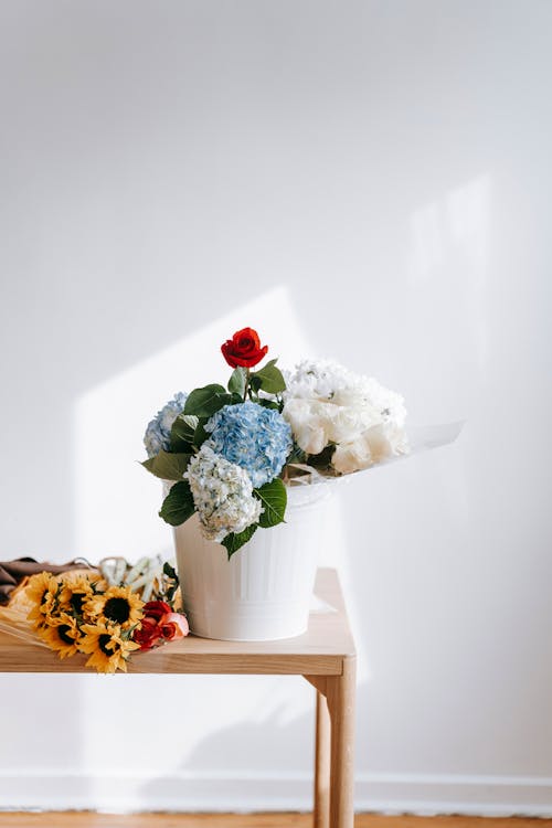 Bouquet with hydrangea flowers and roses in light vase placed on table near sunflowers against white wall in sunlight