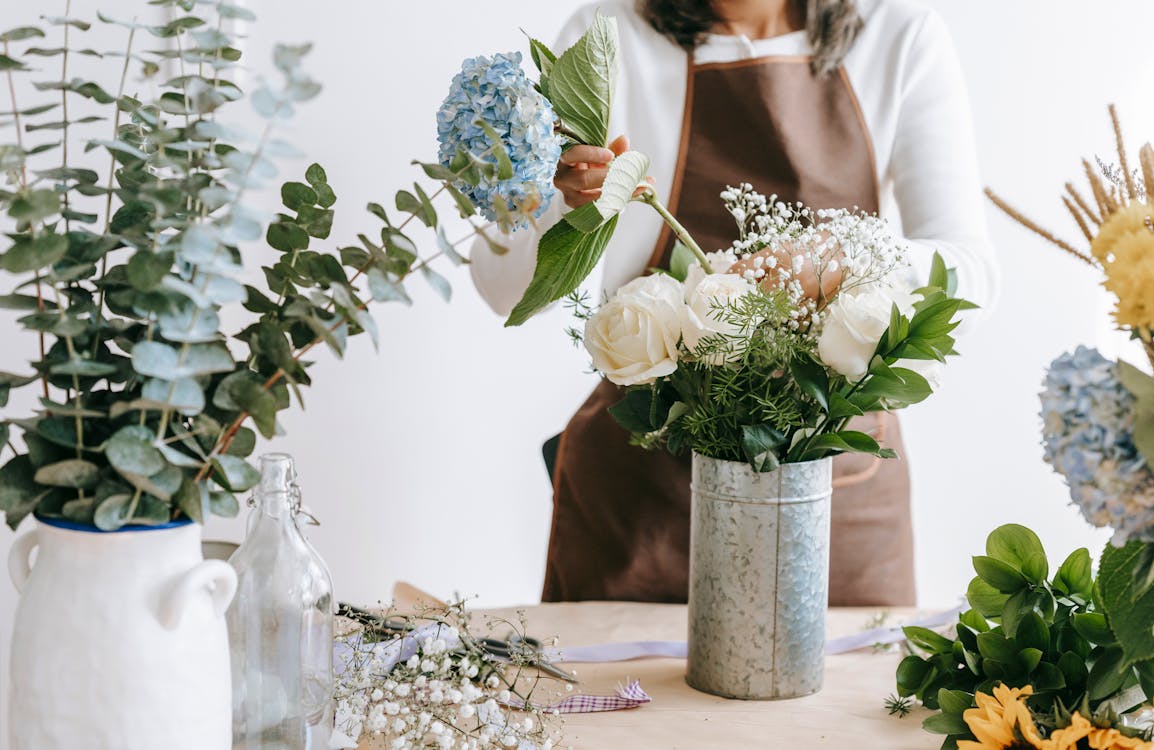 Free Crop unrecognizable female florist arranging hydrangea flowers into vase with roses while standing at table with assorted plants in room Stock Photo