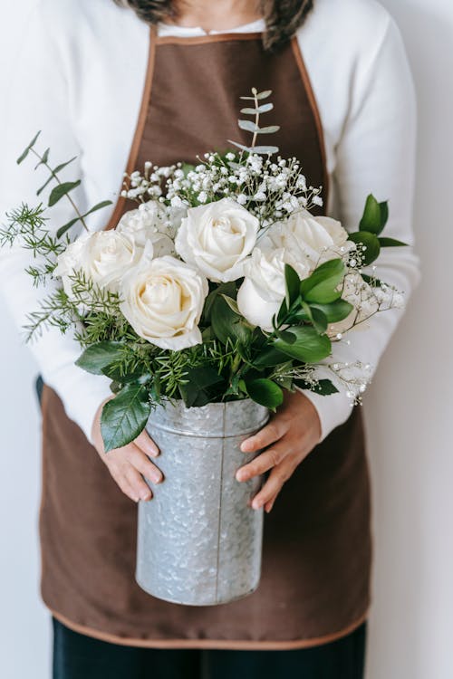 Free Crop unrecognizable female in brown colored apron standing with metal vase with bouquet of white roses in light room Stock Photo