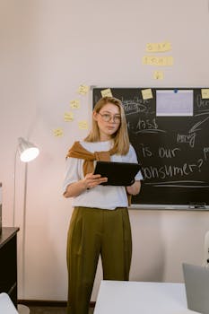 A young woman in business attire stands in an office holding a tablet, ready for a presentation.