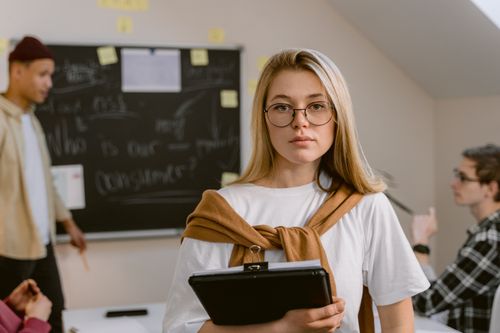 Free A young woman wearing glasses stands in a classroom holding a clipboard. Stock Photo