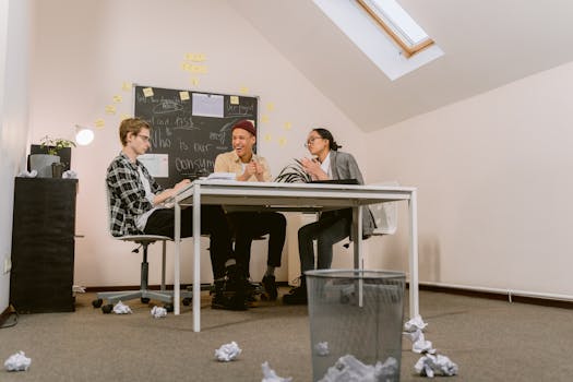 Three diverse adults brainstorm during a meeting with crumpled papers and a blackboard.