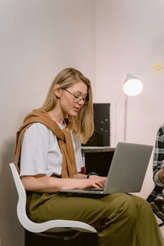 A young woman in casual attire works diligently on her laptop in a modern office setting.