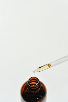 Close-up of a dropper releasing liquid into a glass bottle on a white surface.