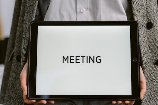 Close-up of a tablet with 'MEETING' on the screen held by a person in a blazer.