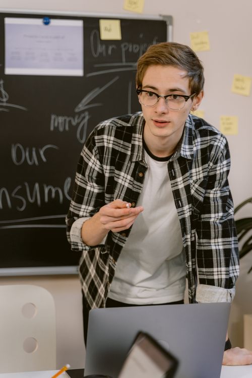 Free Young adult wearing glasses presenting in a classroom with a laptop and blackboard. Stock Photo