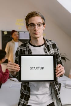 A young professional stands in a startup office holding a digital tablet displaying the word 'STARTUP'.