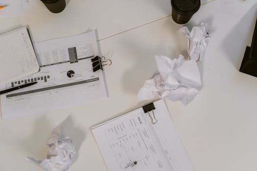 An overhead view of a desk filled with business documents, crumpled paper, and coffee cups.
