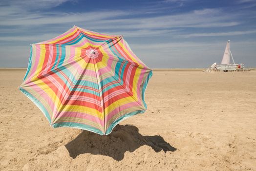 Vibrant striped umbrella on an empty beach with a sailboat in the distance.