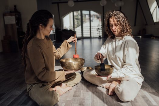 Two women sitting indoors meditating with singing bowls, promoting relaxation and healing.