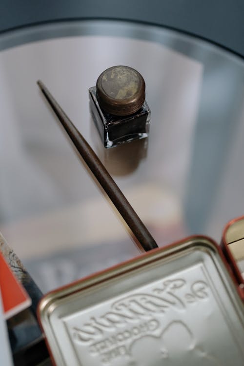 Close-up of an Inkwell and Quill Lying on a Glass Table next to a Metal Box 