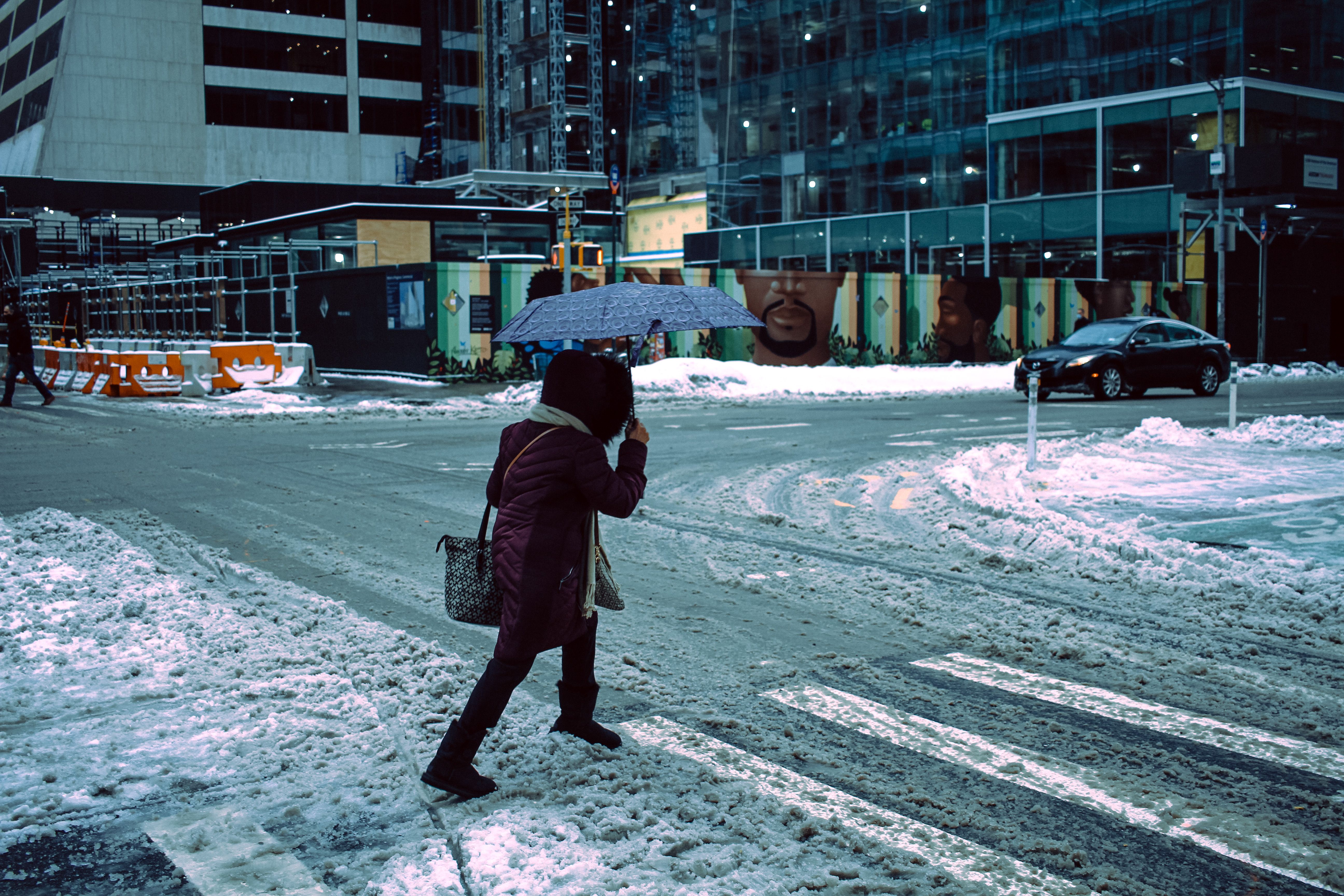 Full body side view of anonymous female with umbrella walking on pedestrian crossing covered with snow on street with building on winter day