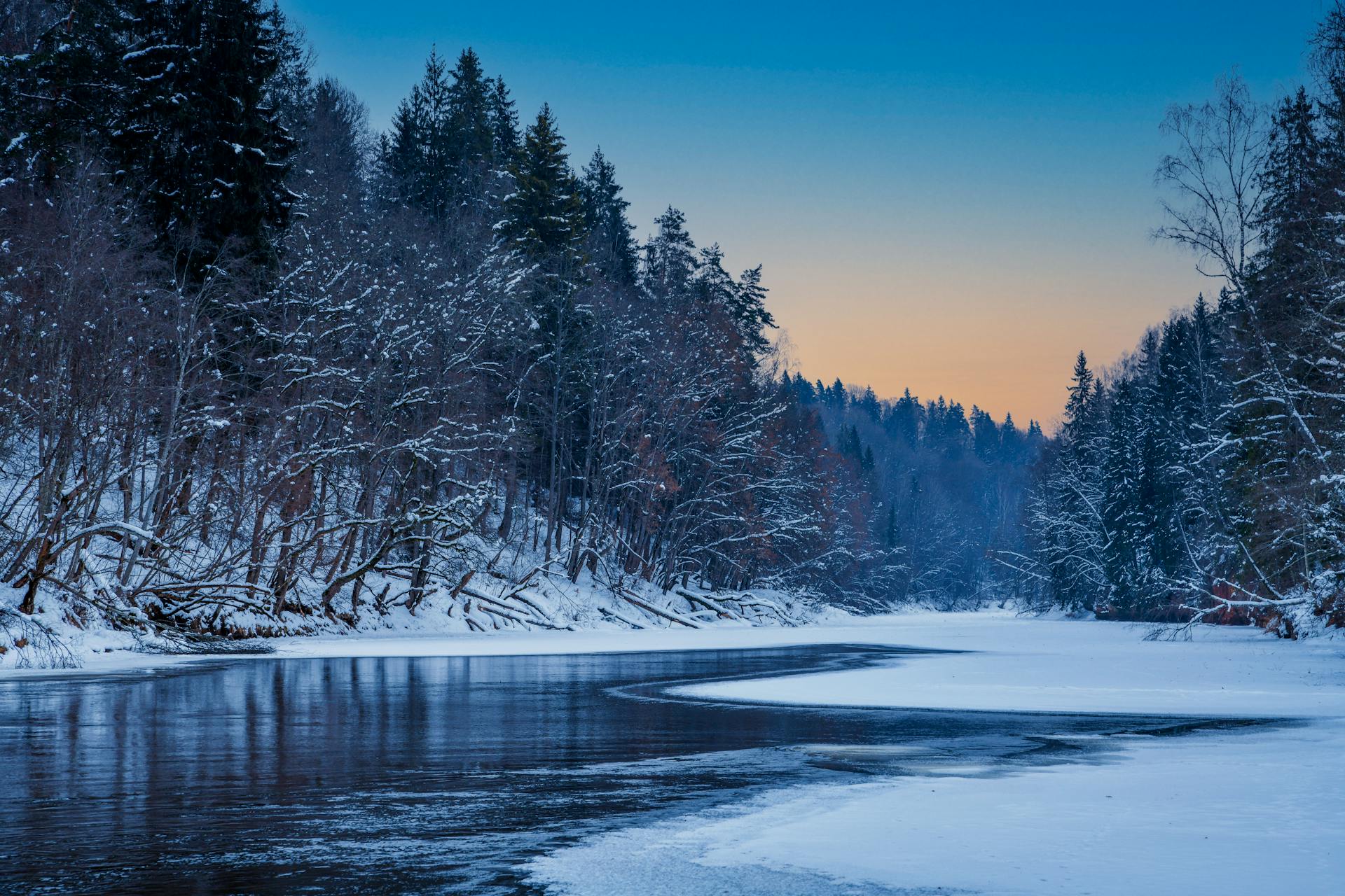 A tranquil winter scene of a snow-covered river in Cēsis, Latvia at dusk.