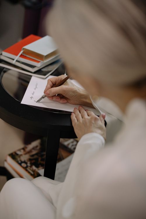 Free A woman writing a personal note on paper with a pen, next to a stack of books. Stock Photo