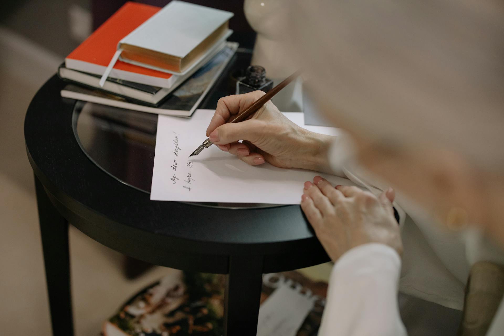 A person writes on white paper using a fountain pen, surrounded by books on a table.