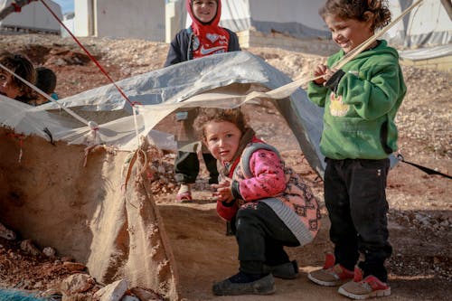 Carefree ethnic preschool children playing in rustic yard with rubbish in settlement of refugee