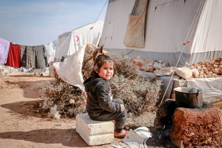 Ethnic Girl Sitting On Concrete Block In Refugee Camp