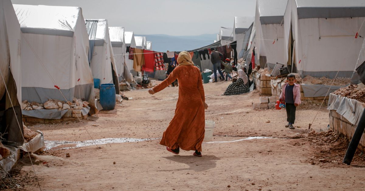 Two young children walk among rows of white refugee tents in a vast, arid desert camp under a clear sky.