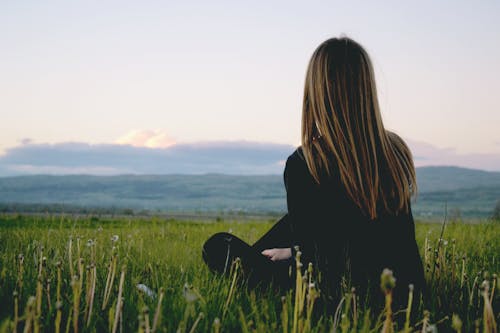 흐린 하늘 아래 산 근처 푸른 잔디 필드에 앉아 검은 긴 소매 셔츠를 입고 여자