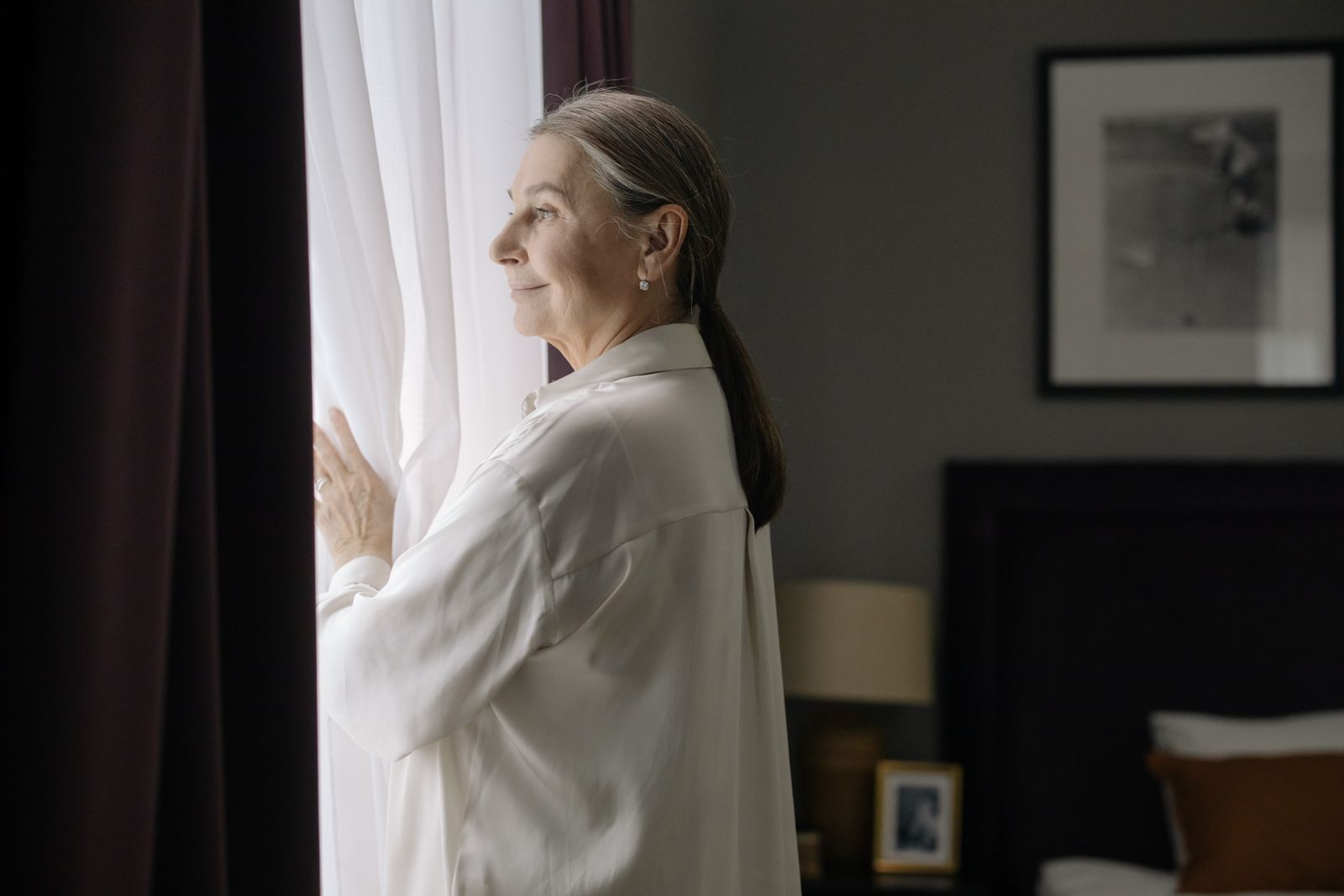 Older woman with gray hair in white shirt smiling softly beside sheer curtains in bedroom window light
