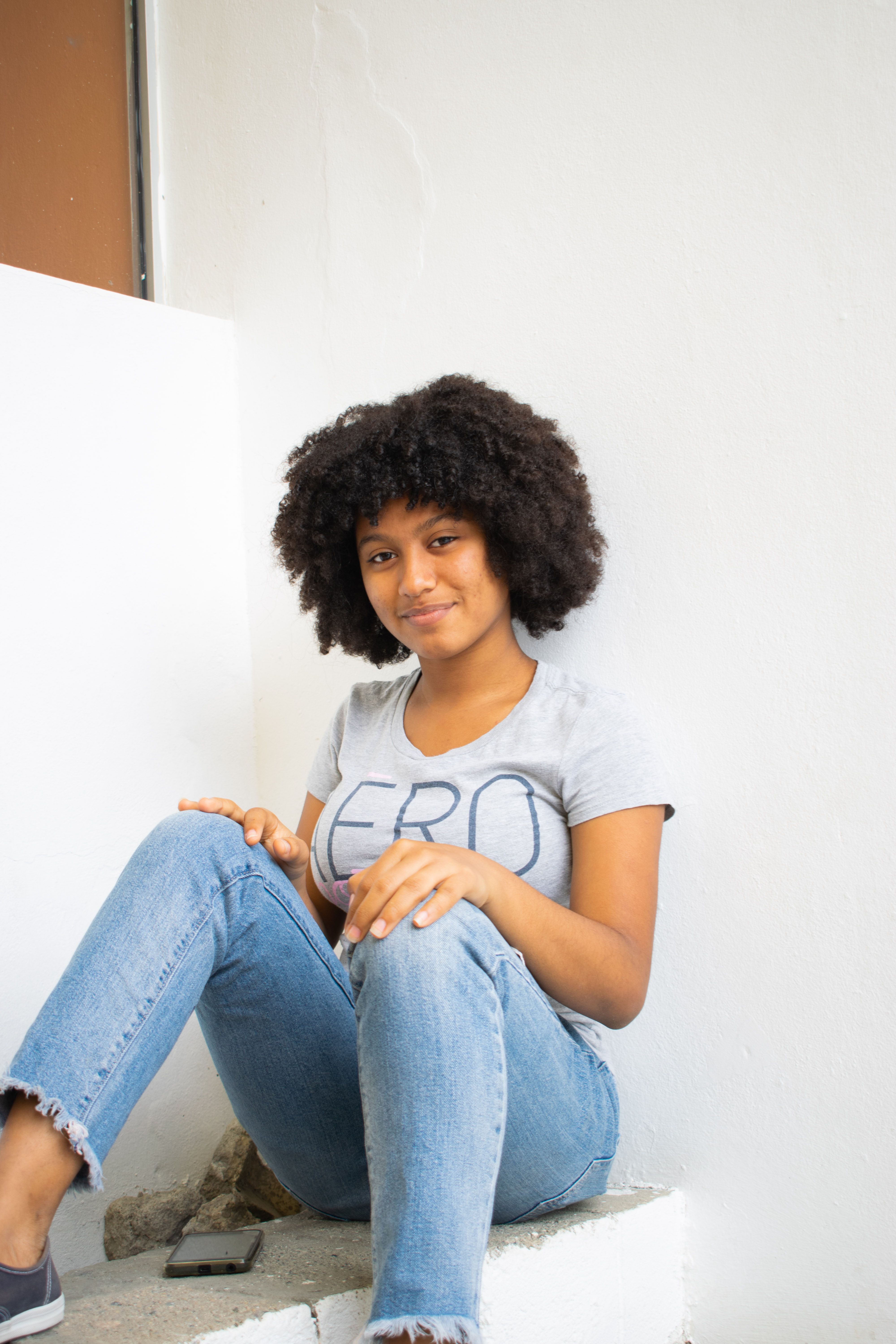 A young woman with afro hair sits indoors, smiling casually in a relaxed pose.