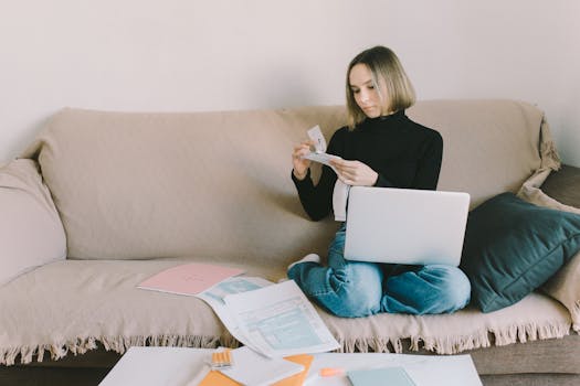 A woman working from home on a sofa with a laptop and documents, managing finances.