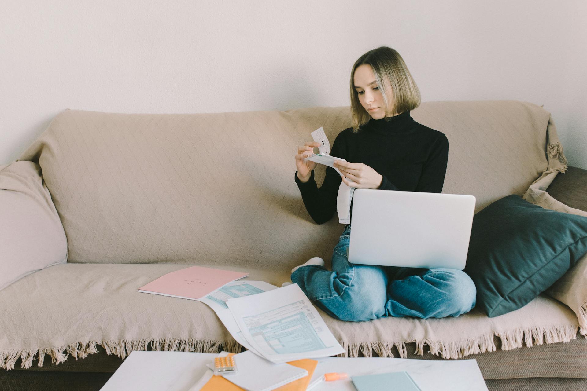 A woman working from home on a sofa with a laptop and documents, managing finances.