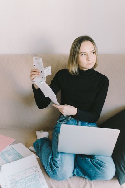 A young woman in casual attire reviewing financial documents with a laptop on a sofa at home.