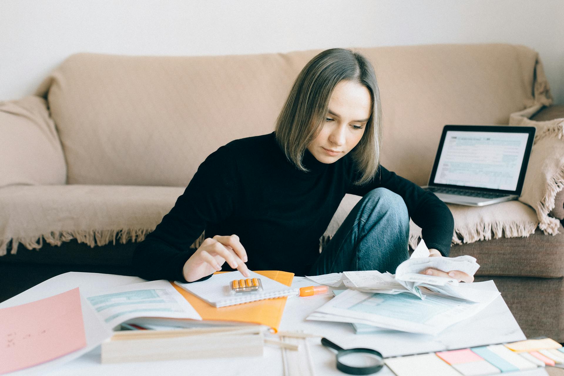 Young woman handling financial tasks with papers and laptop in cozy living room.