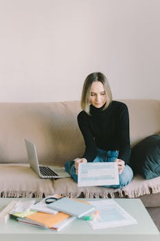 Young woman in a black sweater reviews documents while sitting on a sofa with a laptop nearby.