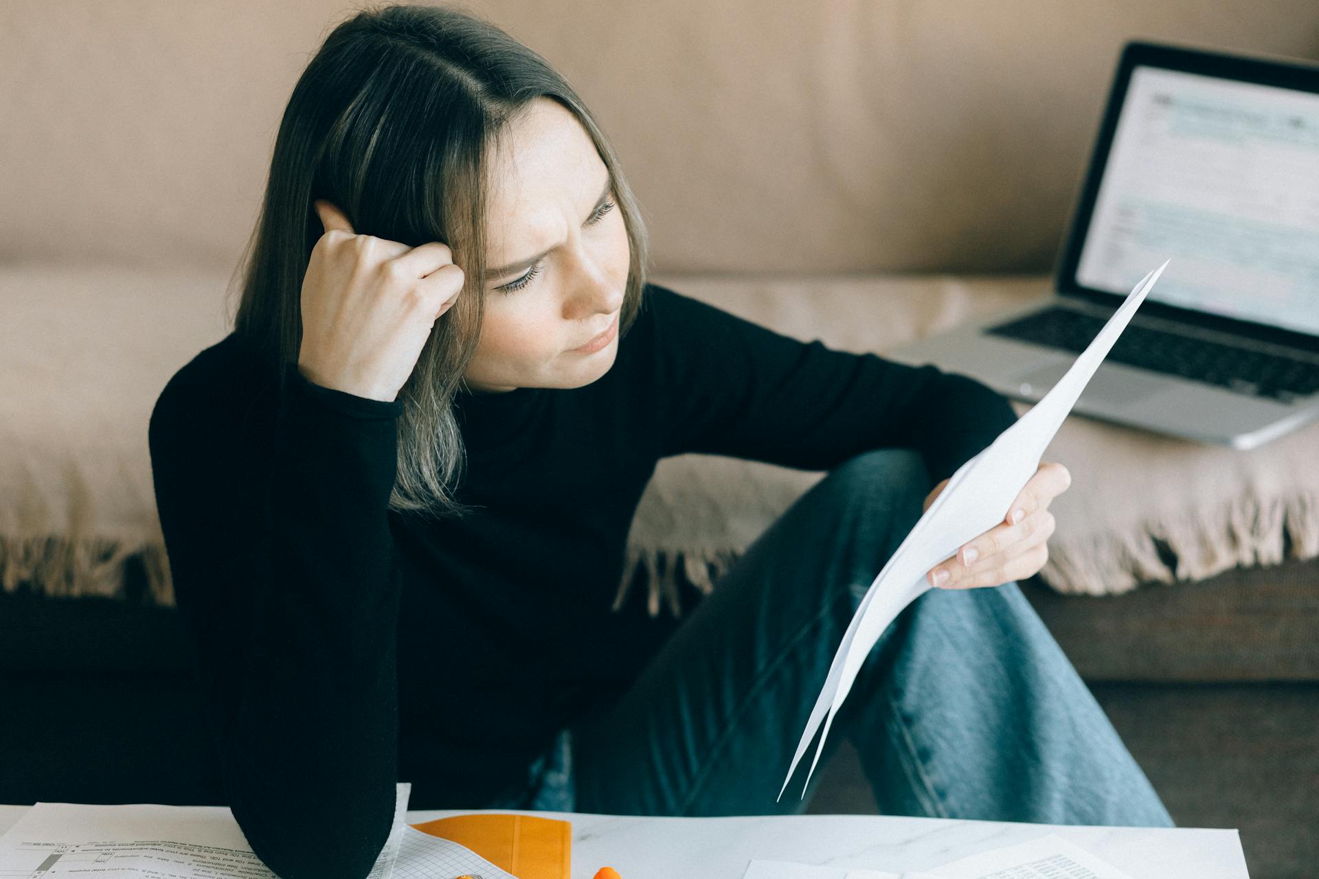 Focused woman analyzing papers with a laptop open, symbolizing thoughtful consideration.