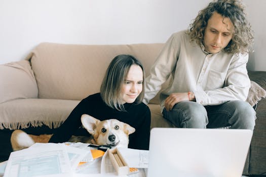 A couple looking at a laptop while sitting on a couch with their pet Corgi amidst paperwork indoors.